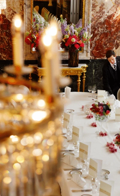 Unter glitzernden Kronleuchtern und prächtigen Marmorwänden bereiten zwei Mitarbeiter in formeller Kleidung im Hotel Sacher Wien sorgfältig einen opulenten Esstisch vor, der mit frischen Blumen und eleganten Gedecken geschmückt ist.