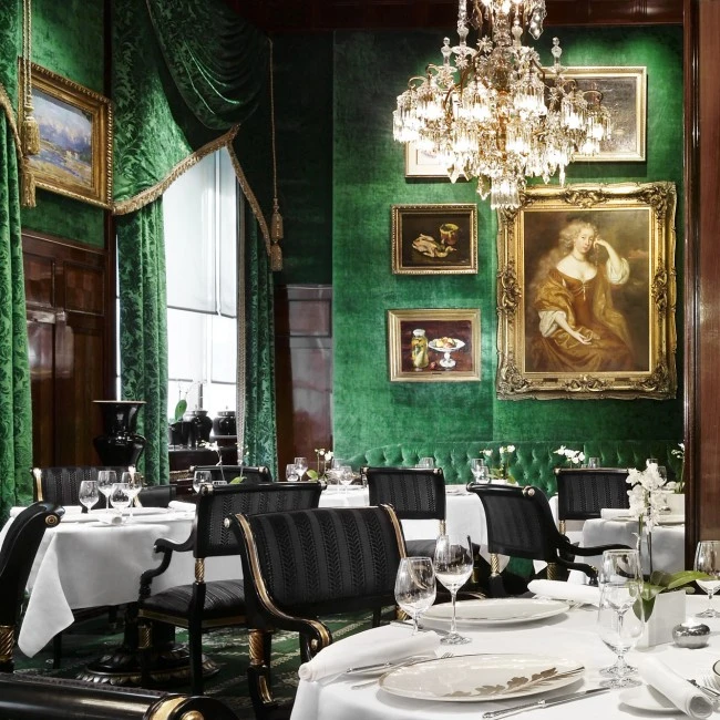 Eleganter Speiseraum mit weißen Tischdecken, goldverziertem Porzellan und Orchideendekor. Die Wände sind in edlem Smaragdgrün gehalten, geschmückt mit klassischen Gemälden in goldenen Rahmen.