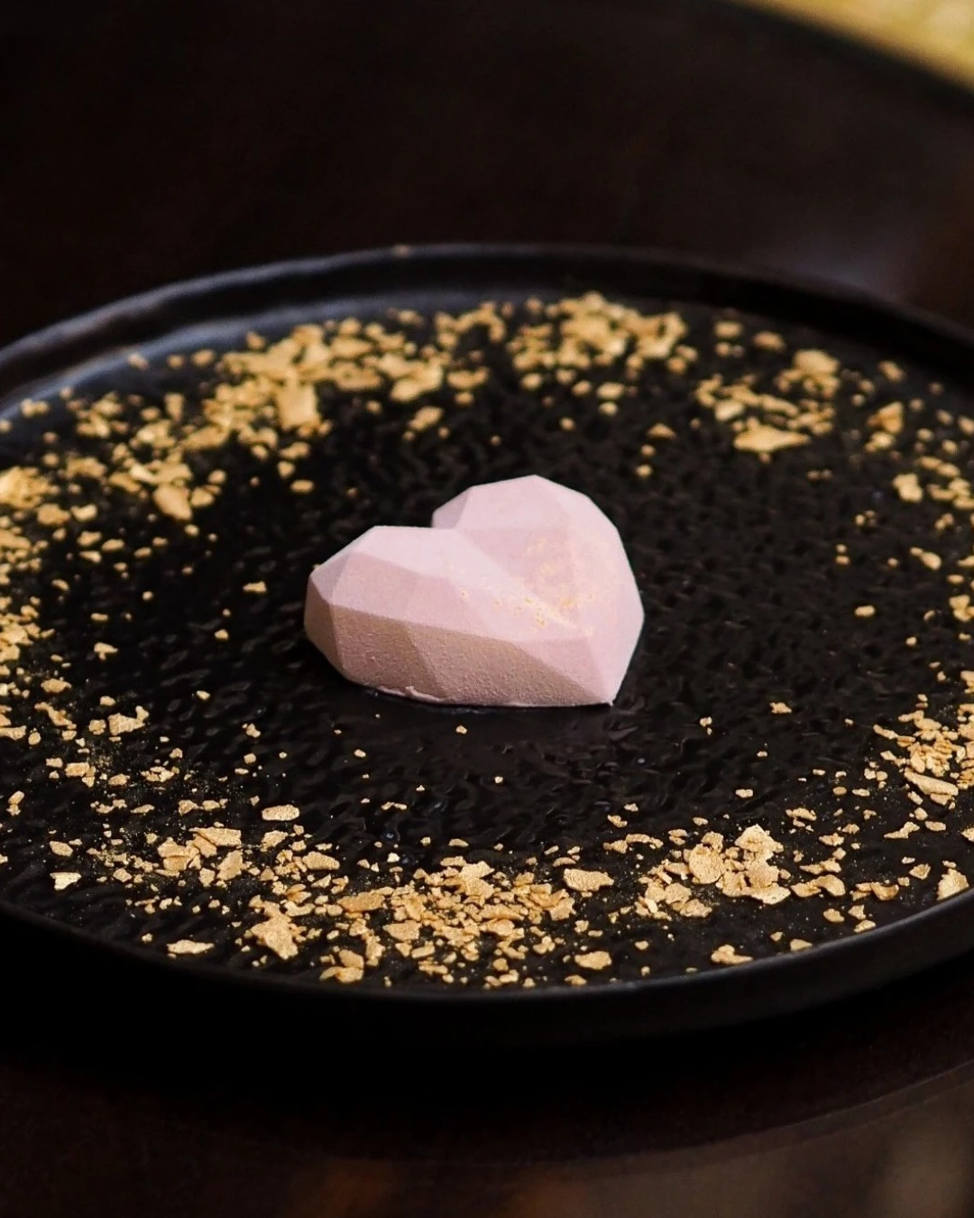Herzförmige, rosafarbene Praline mit geometrischem Muster, präsentiert auf einem schwarzen Teller mit goldglänzenden Streuseln.
