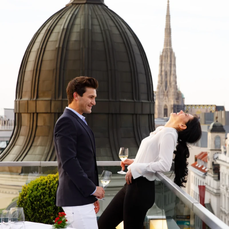 Romantisches Abendessen im Hotel Sacher Wien: Ein Paar genießt Weißwein auf der Dachterrasse mit exklusivem Blick auf den Stephansdom und die Wiener Altstadt.