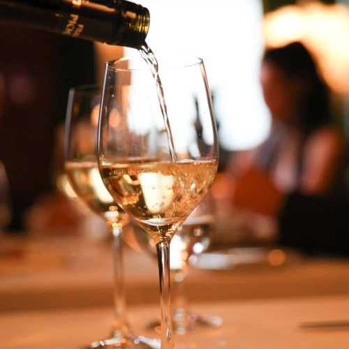 Ein Glas Weißwein wird auf einem elegant gedeckten Tisch eingeschenkt. Im Vordergrund sprudelt der Wein mit feinen Bläschen, im Hintergrund sitzen stilvoll gekleidete Gäste in warmer, verschwommener Beleuchtung