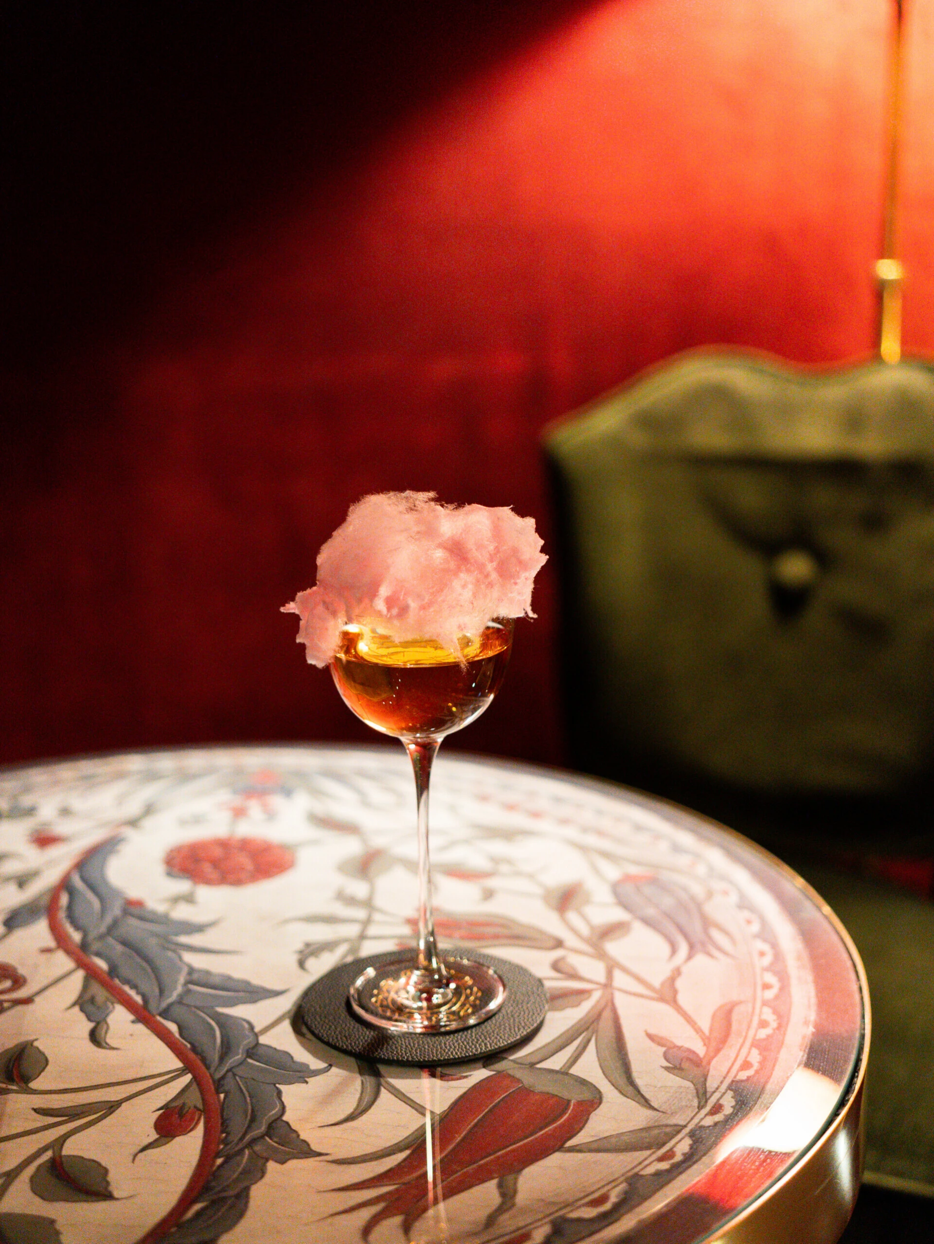 Ein edler bernsteinfarbener Cocktail mit rosa Zuckerwatte-Garnitur auf einem gemusterten Marmortisch.