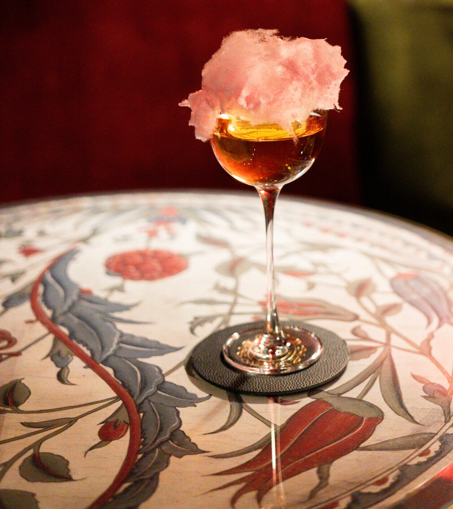 Ein edler bernsteinfarbener Cocktail mit rosa Zuckerwatte-Garnitur auf einem gemusterten Marmortisch.