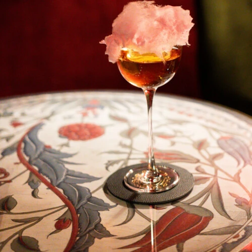 Ein edler bernsteinfarbener Cocktail mit rosa Zuckerwatte-Garnitur auf einem gemusterten Marmortisch.