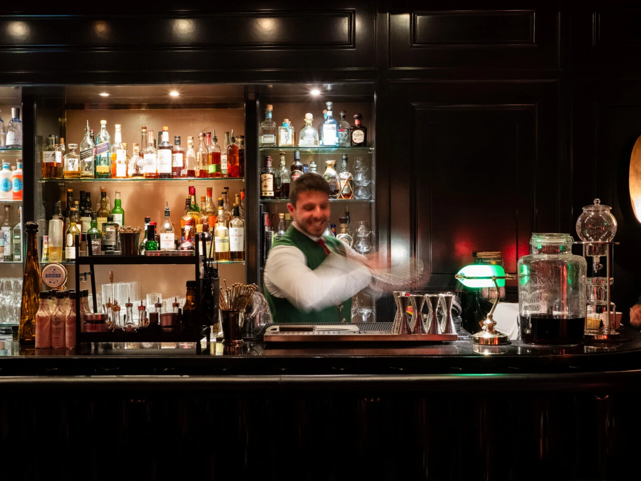 Professioneller Barkeeper in grüner Weste beim Mixen eines Cocktails hinter einer gut sortierten Hotelbar.