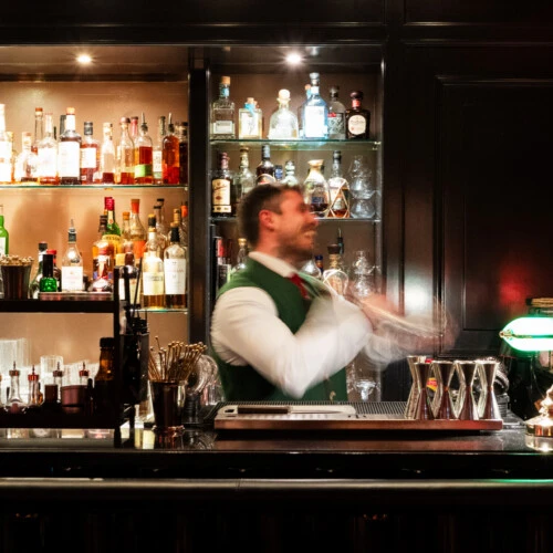 Professioneller Barkeeper in grüner Weste beim Mixen eines Cocktails hinter einer gut sortierten Hotelbar.