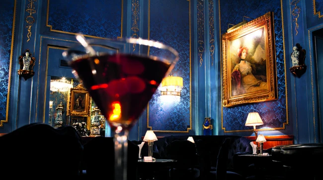 Ein eleganter Salon in tiefem Blau mit goldenen Zierleisten und floraler Tapete. Im Mittelpunkt steht ein Gemälde einer Dame im Goldrahmen, beleuchtet von Wand- und Tischlampen mit Stoffschirmen.