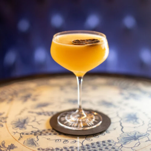 Cocktail in einer Coupette mit getrockneter Zitrusgarnitur auf einem Untersetzer auf einem dekorativen Tisch.