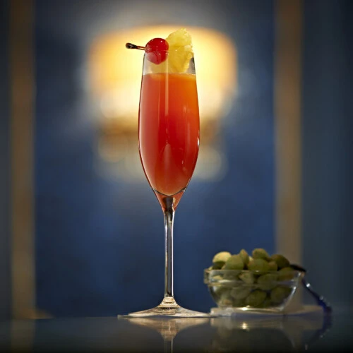 Hoher Cocktail im schlanken Champagnerglas mit rot-orangem Farbverlauf, garniert mit Ananasstück und Cocktailkirsche auf einem Spieß