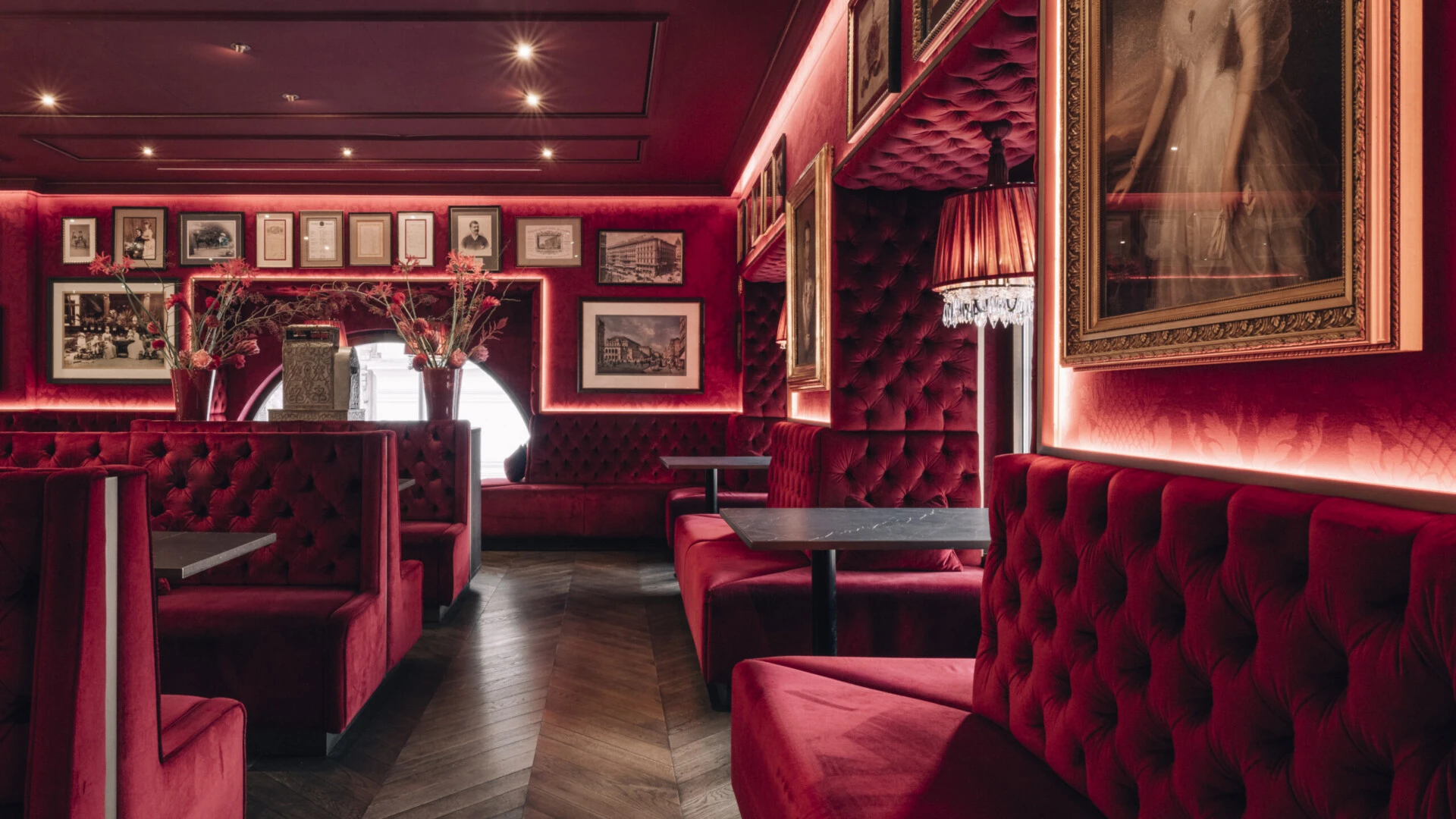 Sitzbänke mit rotem Samtbezug und Rautensteppung, dunkler Fischgrätparkettboden, schwarze Tische, rote Wände mit gerahmten Porträts und Fotografien, eingelassene Beleuchtung, Kristalllampen mit roten Schirmen.