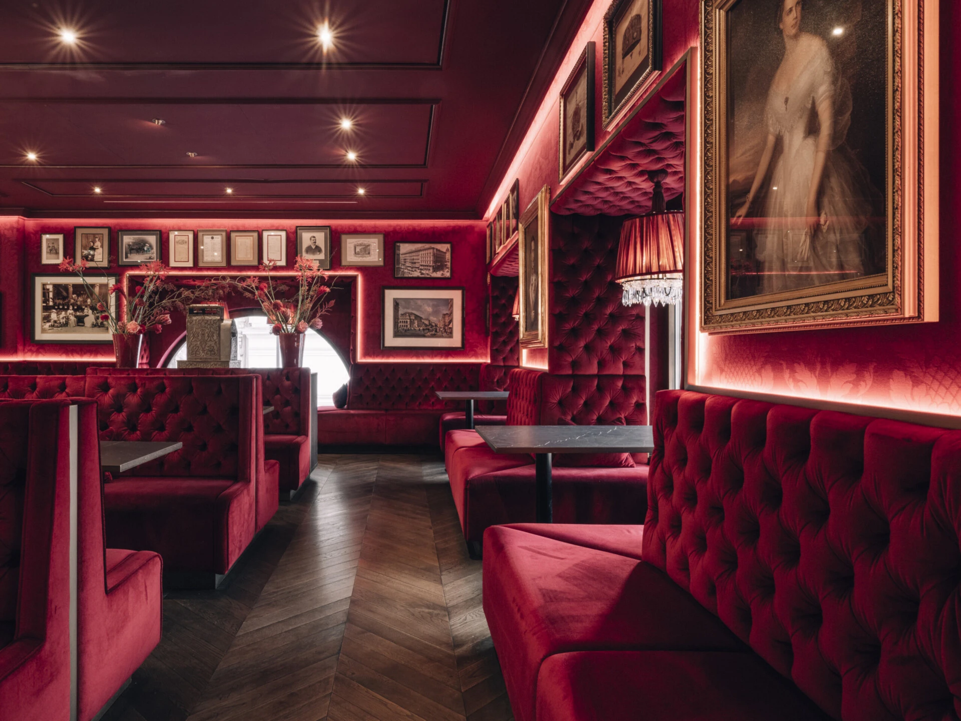 Sitzbänke mit rotem Samtbezug und Rautensteppung, dunkler Fischgrätparkettboden, schwarze Tische, rote Wände mit gerahmten Porträts und Fotografien, eingelassene Beleuchtung, Kristalllampen mit roten Schirmen.