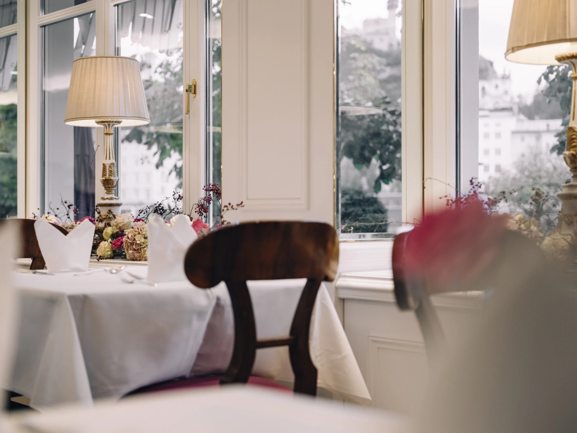 Ein gedeckter Tisch mit roten Polsterstühlen steht in einem hellen Raum am Fenster. Auf der Fensterbank liegen Blumenarrangements, daneben steht eine klassische Tischlampe.