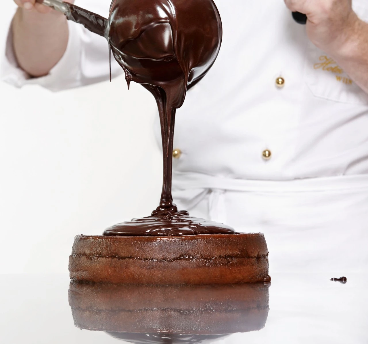 Ein Konditor in weißer Uniform gießt glänzende Schokoladenglasur aus einer Kelle über eine Original Sacher-Torte.