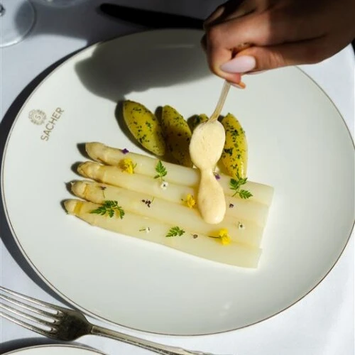 Spargelgericht auf einem Sacher-Teller, serviert mit Kartoffeln und Sauce, während eine Hand das Gericht anrichtet