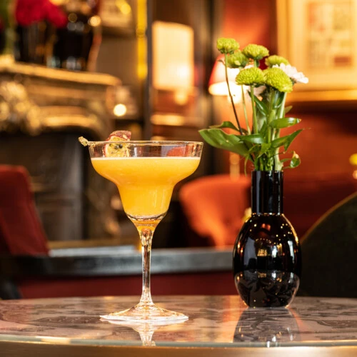 Leuchtend gelber Cocktail in eleganter Coupette mit Passionsfruchtscheibe, stilvoll serviert auf einem Marmortisch neben einer schwarzen Vase mit frischen Blumen, vor gemütlich warmem Barambiente.