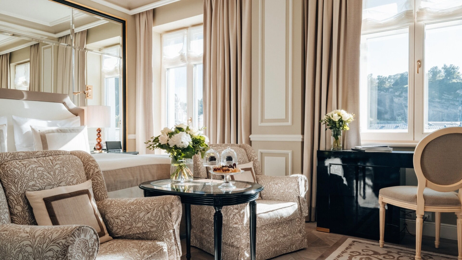 Deluxe Zimmer mit Ausblick im Hotel Sacher Salzburg, mit gemusterten Sesseln, frischen Blumen, kleinen Desserts und elegantem Bett im Hintergrund