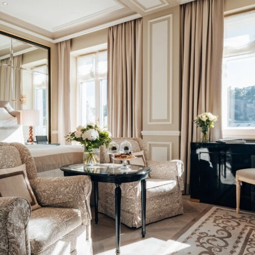 Deluxe Zimmer mit Ausblick im Hotel Sacher Salzburg, mit gemusterten Sesseln, frischen Blumen, kleinen Desserts und elegantem Bett im Hintergrund