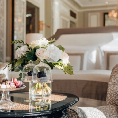 Detailaufnahme des Glastischs im Deluxe Zimmer mit Ausblick im Hotel Sacher Salzburg, mit Blumenstrauß, kleinen Desserts und elegantem Bett im Hintergrund