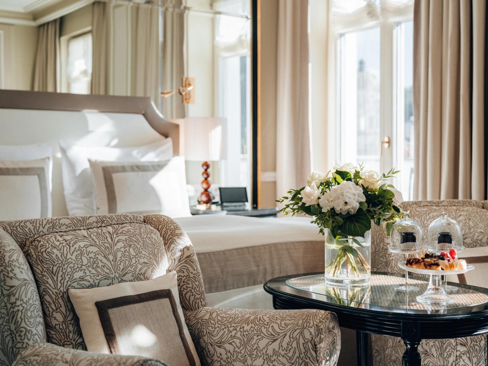 Sitzbereich im Deluxe Zimmer mit Ausblick im Hotel Sacher Salzburg, mit gemusterten Sesseln, frischen Blumen, kleinen Desserts und elegantem Bett im Hintergrund