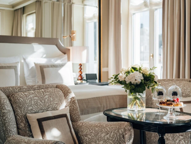 Sitzbereich im Deluxe Zimmer mit Ausblick im Hotel Sacher Salzburg, mit gemusterten Sesseln, frischen Blumen, kleinen Desserts und elegantem Bett im Hintergrund
