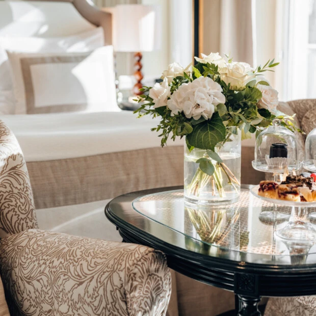 Stilvoll gedeckter Beistelltisch mit frischen weißen Rosen und Hortensien in einer Vase sowie einer Etagere mit kleinen Desserts, eingerahmt von zwei eleganten Polstersesseln vor bodenlangen Vorhängen.