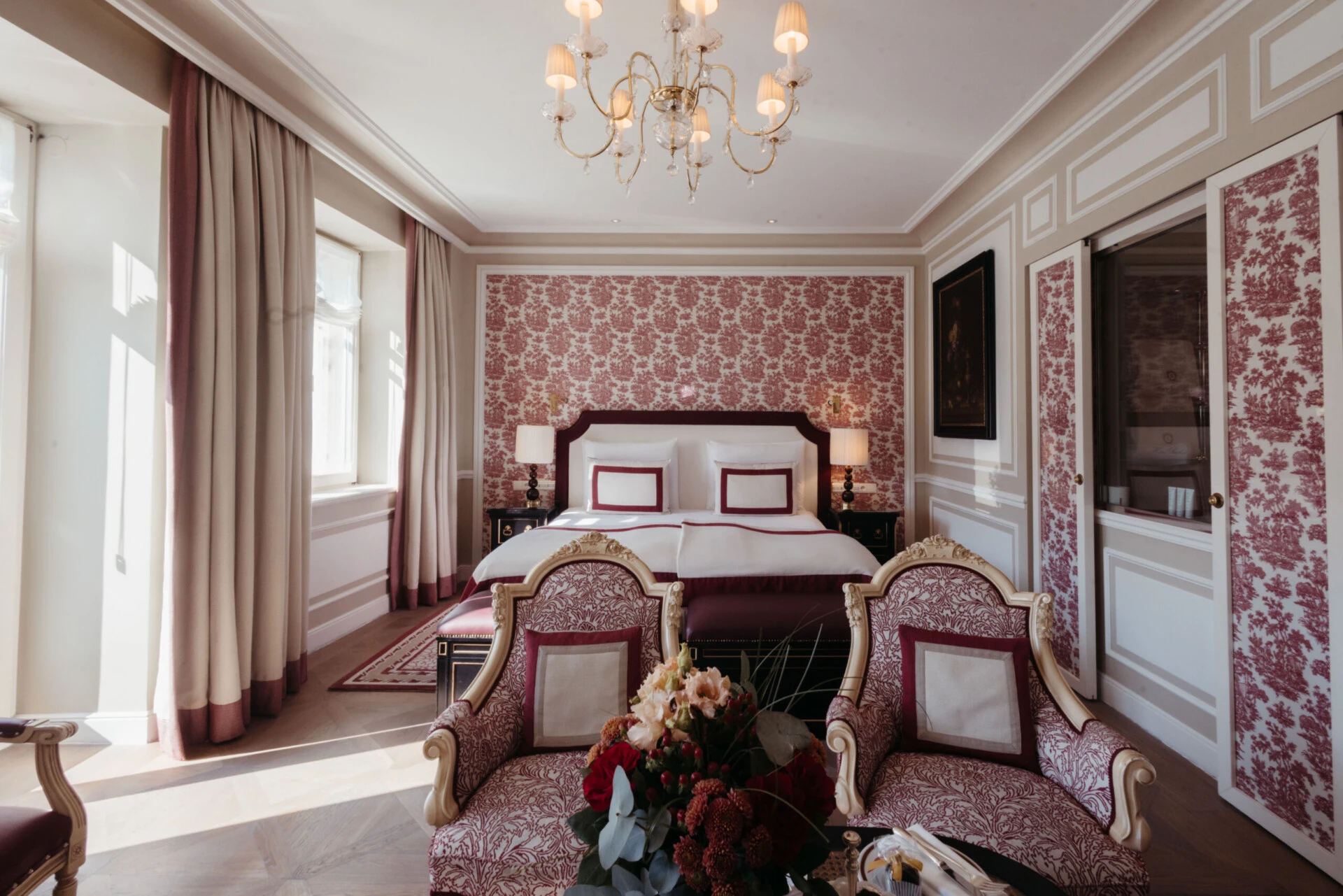 Junior Suite mit Ausblick im Hotel Sacher Salzburg, mit elegantem Bett, roten Mustertapeten, klassischen Sesseln und Kristalllüster
