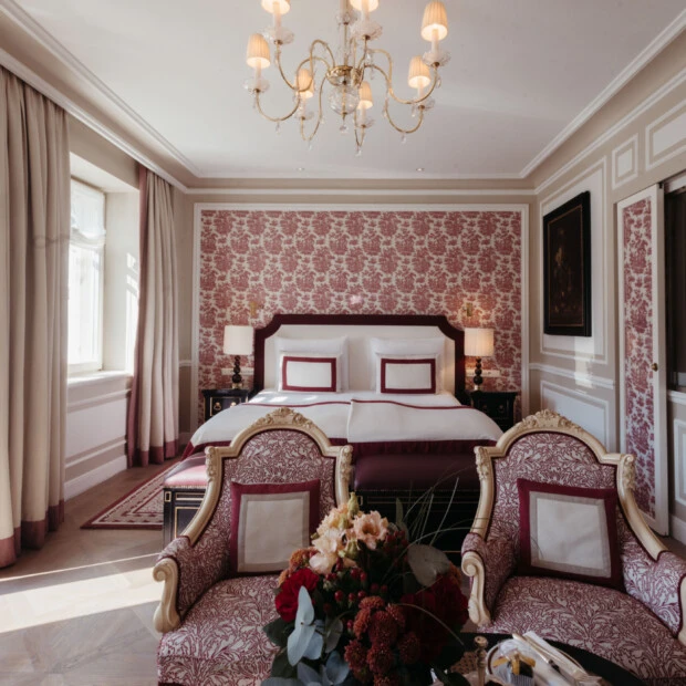Junior Suite mit Ausblick im Hotel Sacher Salzburg, mit elegantem Bett, roten Mustertapeten, klassischen Sesseln und Kristalllüster