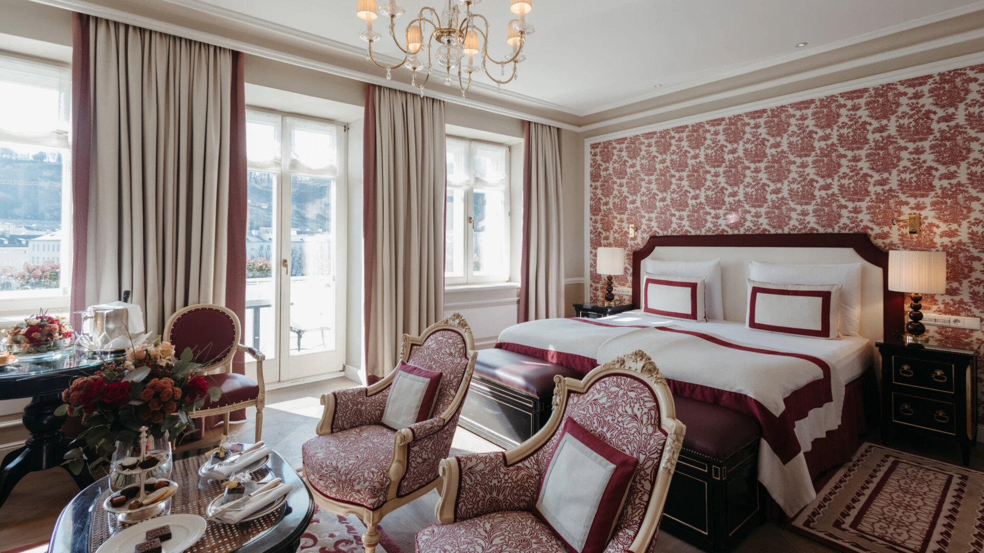 Junior Suite mit Ausblick im Hotel Sacher Salzburg, mit elegantem Doppelbett, klassischen Sesseln, roten Musterdetails und großen Fenstern