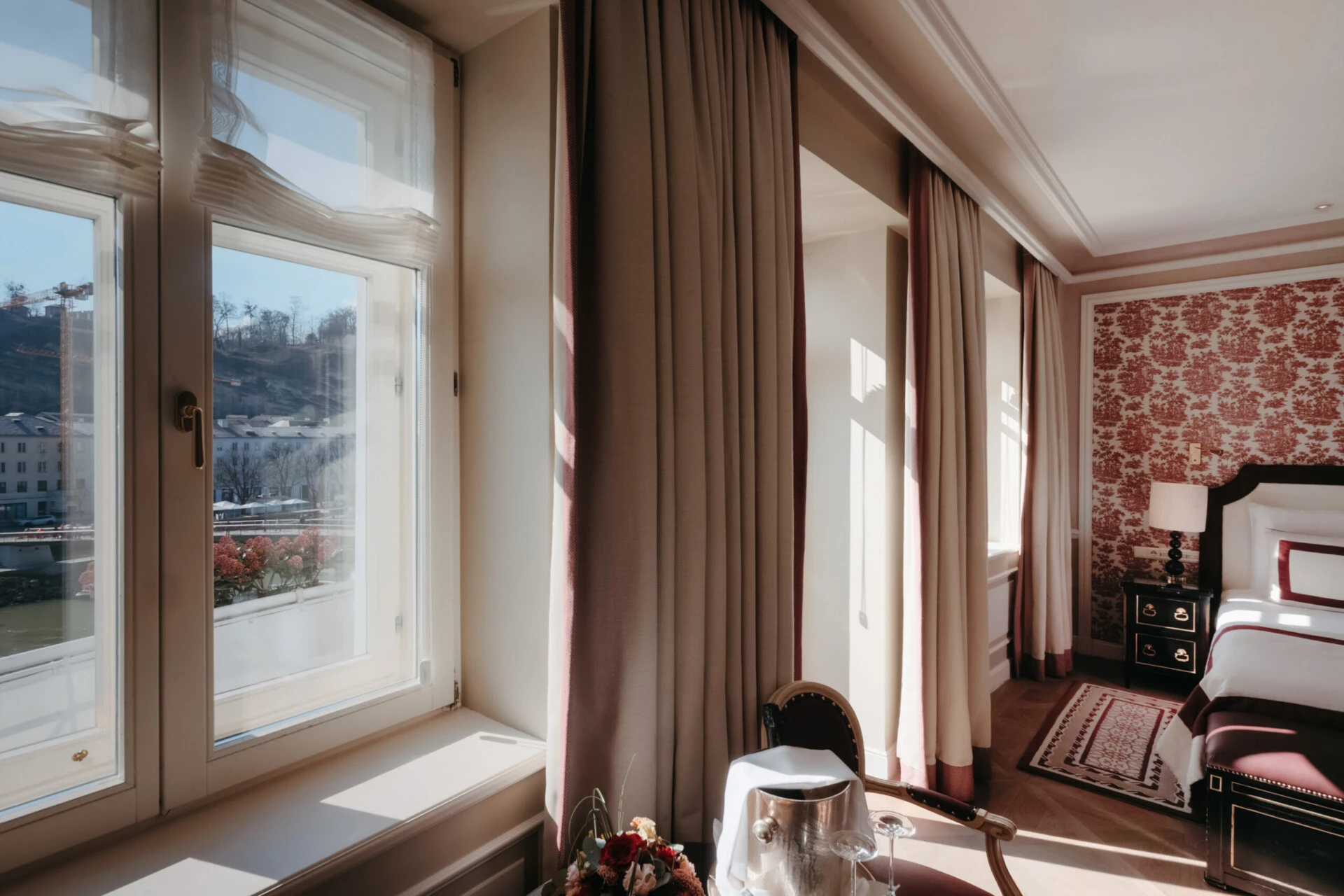 Junior Suite mit Ausblick im Hotel Sacher Salzburg, mit großen Fenstern, Blick auf die Salzach und elegantem Bett mit roten Designdetails