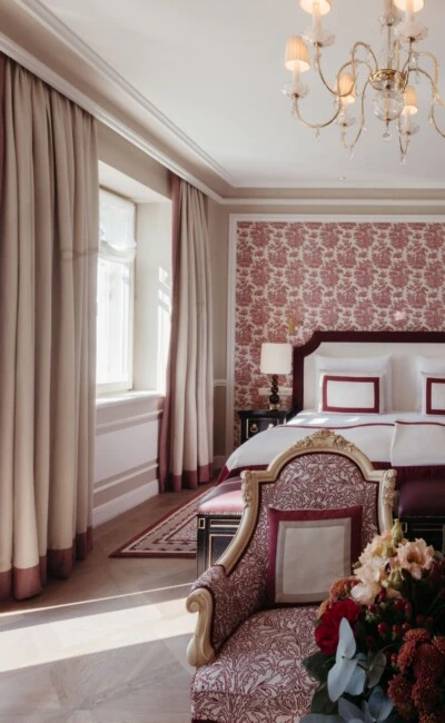 Junior Suite mit Ausblick im Hotel Sacher Salzburg, mit elegantem Bett, roten Mustertapeten, klassischen Sesseln und Kristalllüster