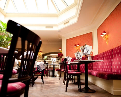 Heller, einladender Caféraum mit Tageslicht durch ein großzügiges Oberlicht, eleganter roter Wandbespannung, samtgepolsterten Sitzbänken und klassisch dunklem Holzmobiliar