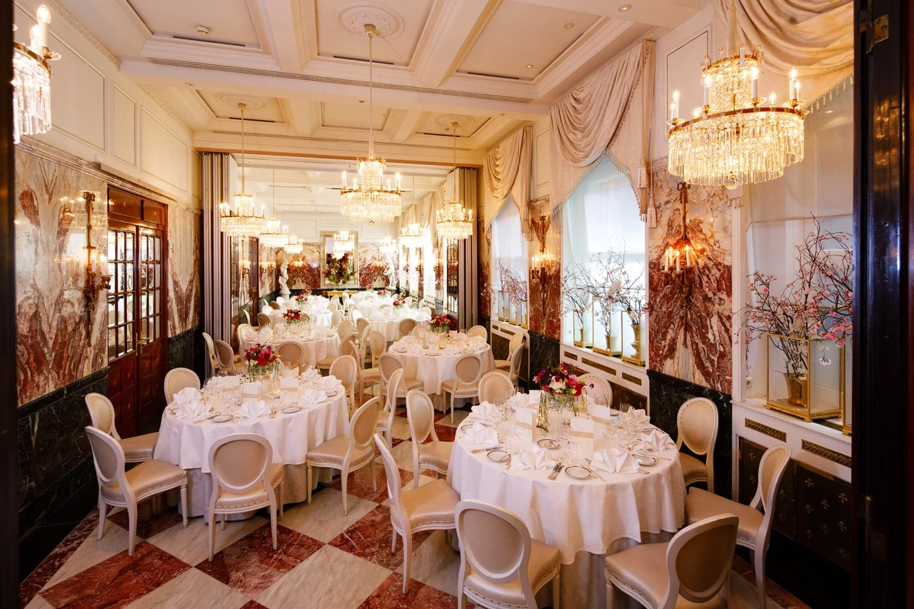 Prunkvoll gedeckter Festsaal mit runden, elegant eingedeckten Tischen, umgeben von klassisch gepolsterten Stühlen, reich verzierten Kronleuchtern und floralen Akzenten