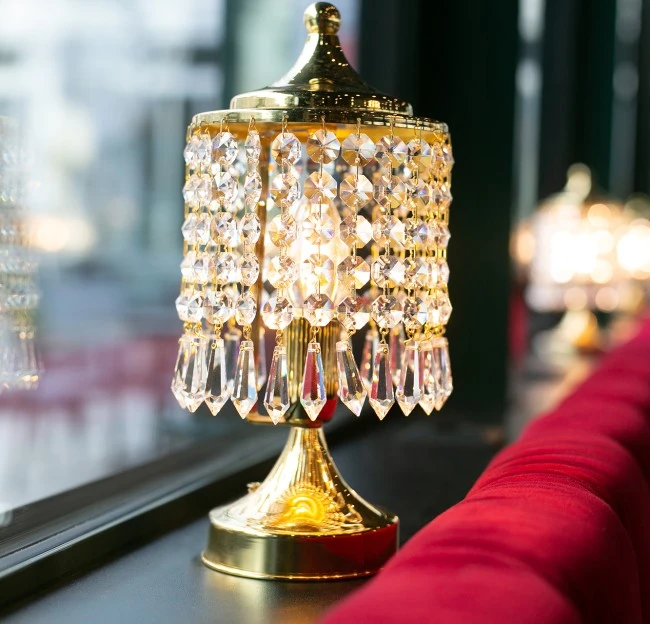 Eine elegante Kristalllampe mit goldfarbenem Fuß und funkelnden Prismen steht auf einer Fensterbank vor rotem Samtpolster, strahlt warmes Licht aus und verleiht dem Raum luxuriöse Atmosphäre.