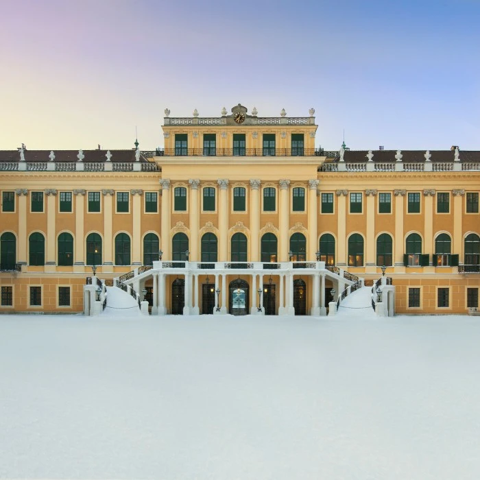 Innenhofansicht von Schloss Schönbrunn im Winter mit verschneitem Vorplatz, symmetrisch gegliederter gelber Fassade, grünen Fensterläden und doppelläufiger Freitreppe in der Mitte; über dem Eingang ein Uhrenturm, umgeben von Statuen.