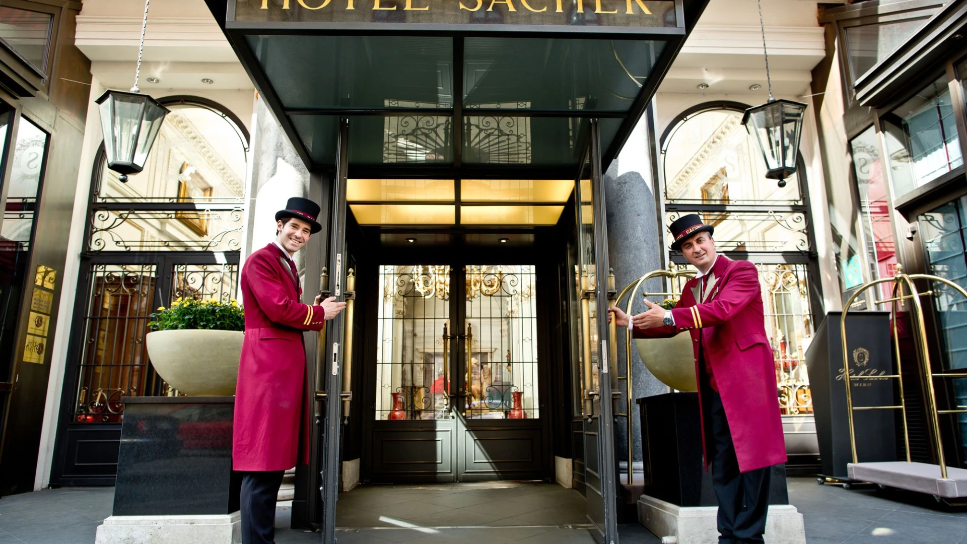 Zwei freundliche Portiers in bordeauxroten Uniformen begrüßen die Gäste mit offenen Türen am Eingang des traditionsreichen Hotel Sacher. Die elegante Glasüberdachung, klassische Laternen und die goldene Schrift verleihen dem Szenario eine stilvolle und einladende Atmosphäre.
