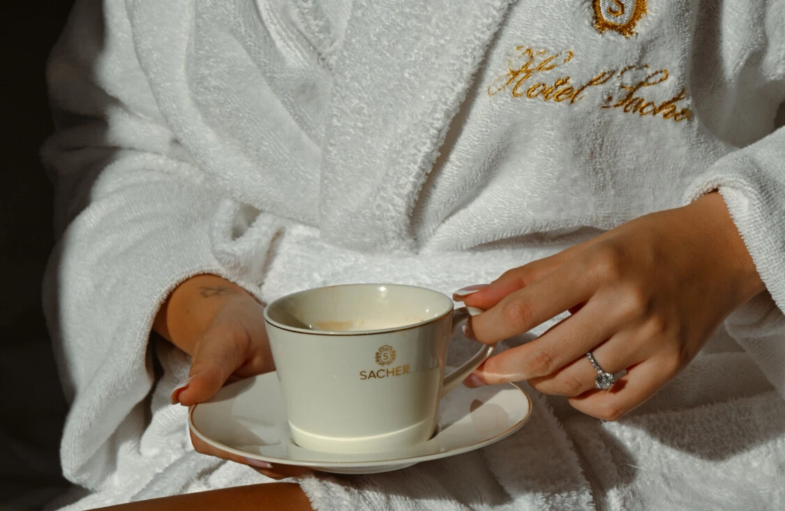Detailaufnahme eines Gastes im Sacher-Bademantel mit Sacher-Tasse im warmen Sonnenlicht.