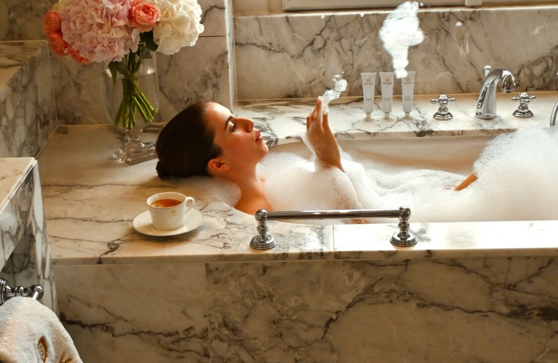 Gast entspannt in einem Schaumbad im eleganten Marmorbad des Hotel Sacher Wien mit Blumen und Sacher-Hausschuhen.
