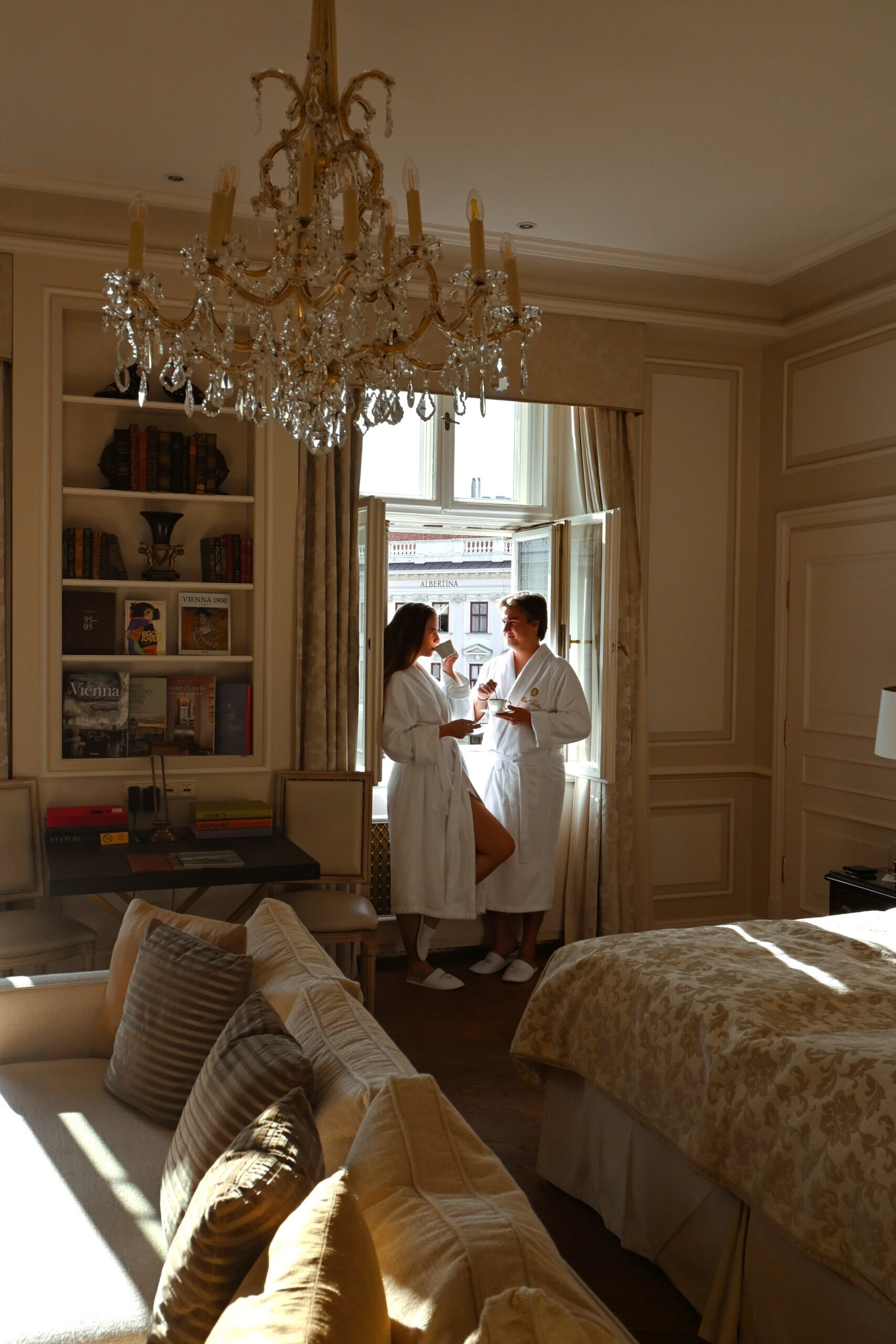Zwei Gäste im Sacher-Bademantel genießen Kaffee am offenen Fenster eines eleganten Zimmers im Hotel Sacher Wien mit Blick auf die Albertina.