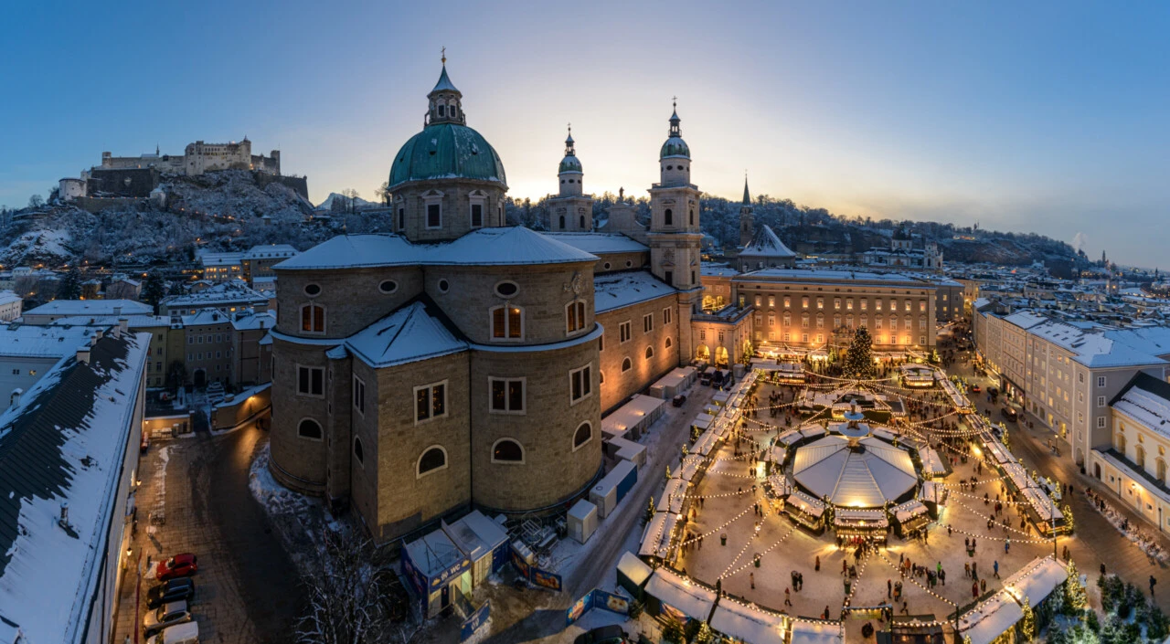 Festlich beleuchteter Weihnachtsmarkt in der verschneiten Salzburger Altstadt mit Salzburger Dom und Festung Hohensalzburg im Hintergrund.