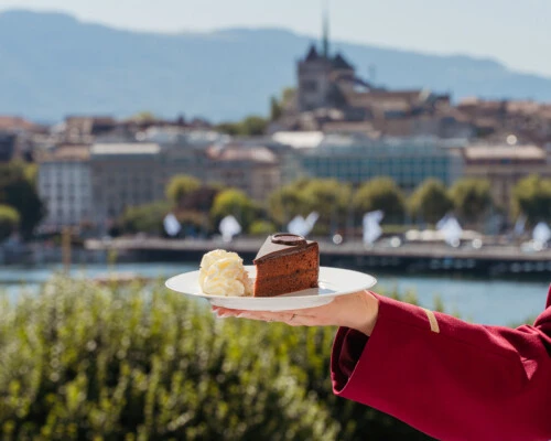 Ein Stück Original Sacher-Torte mit Schlagobers auf einem Teller, gehalten vor einer sonnigen Stadt- und Uferkulisse.