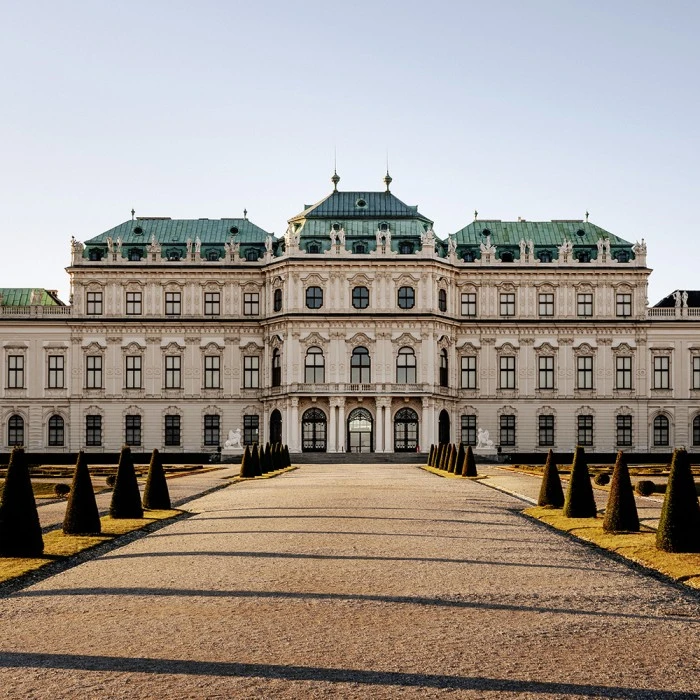 Frontansicht des Oberen Belvedere in Wien bei tiefstehender Sonne, mit symmetrisch angelegtem barocken Garten, kegelförmig beschnittenen Buchsbäumen und klassizistischer Architektur mit grünem Kupferdach und kunstvoll dekorierter Fassade.