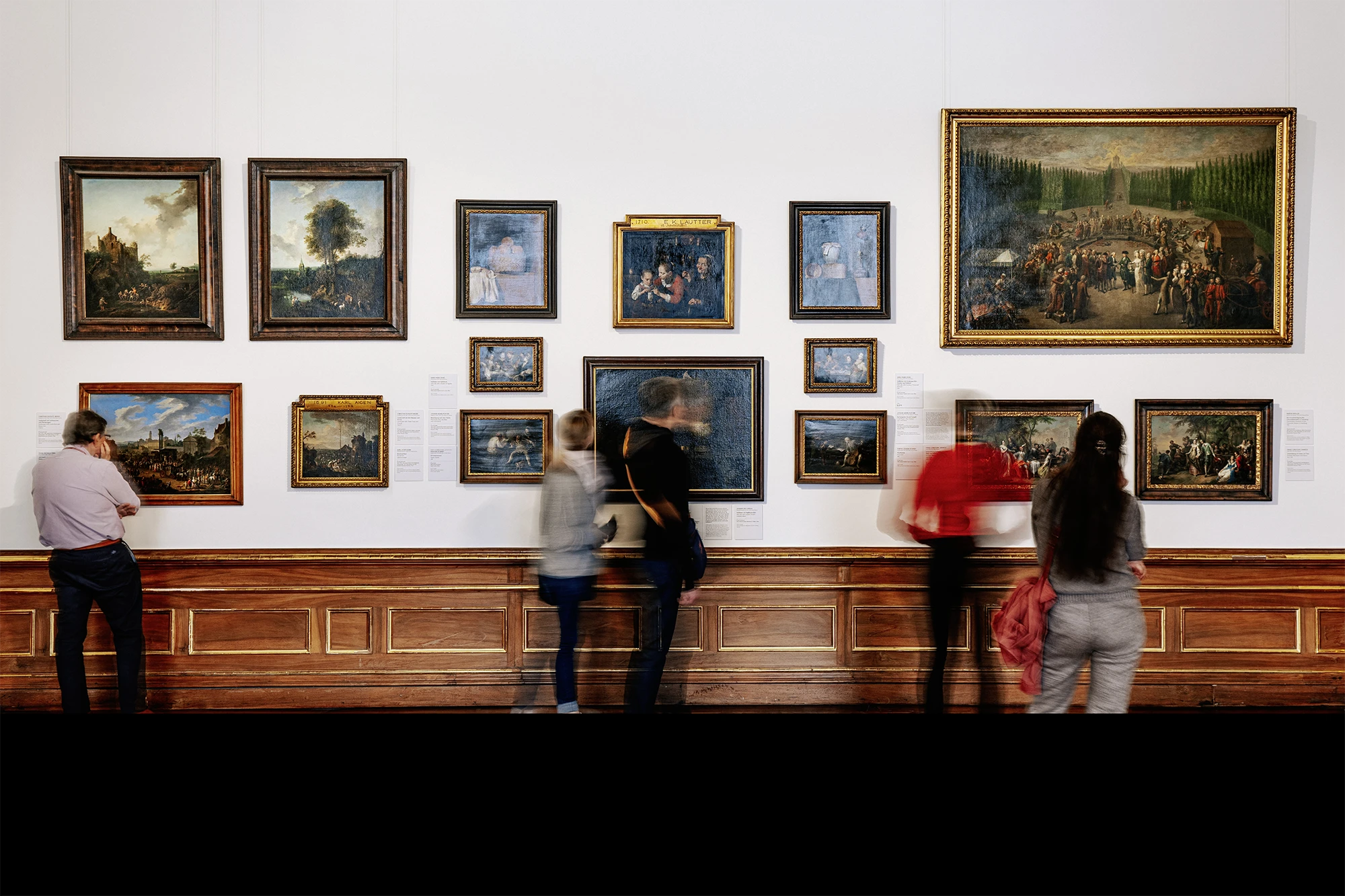 Museumssaal mit weißer Wand und Holzvertäfelung, an der zahlreiche gerahmte Gemälde in verschiedenen Größen dicht gehängt sind