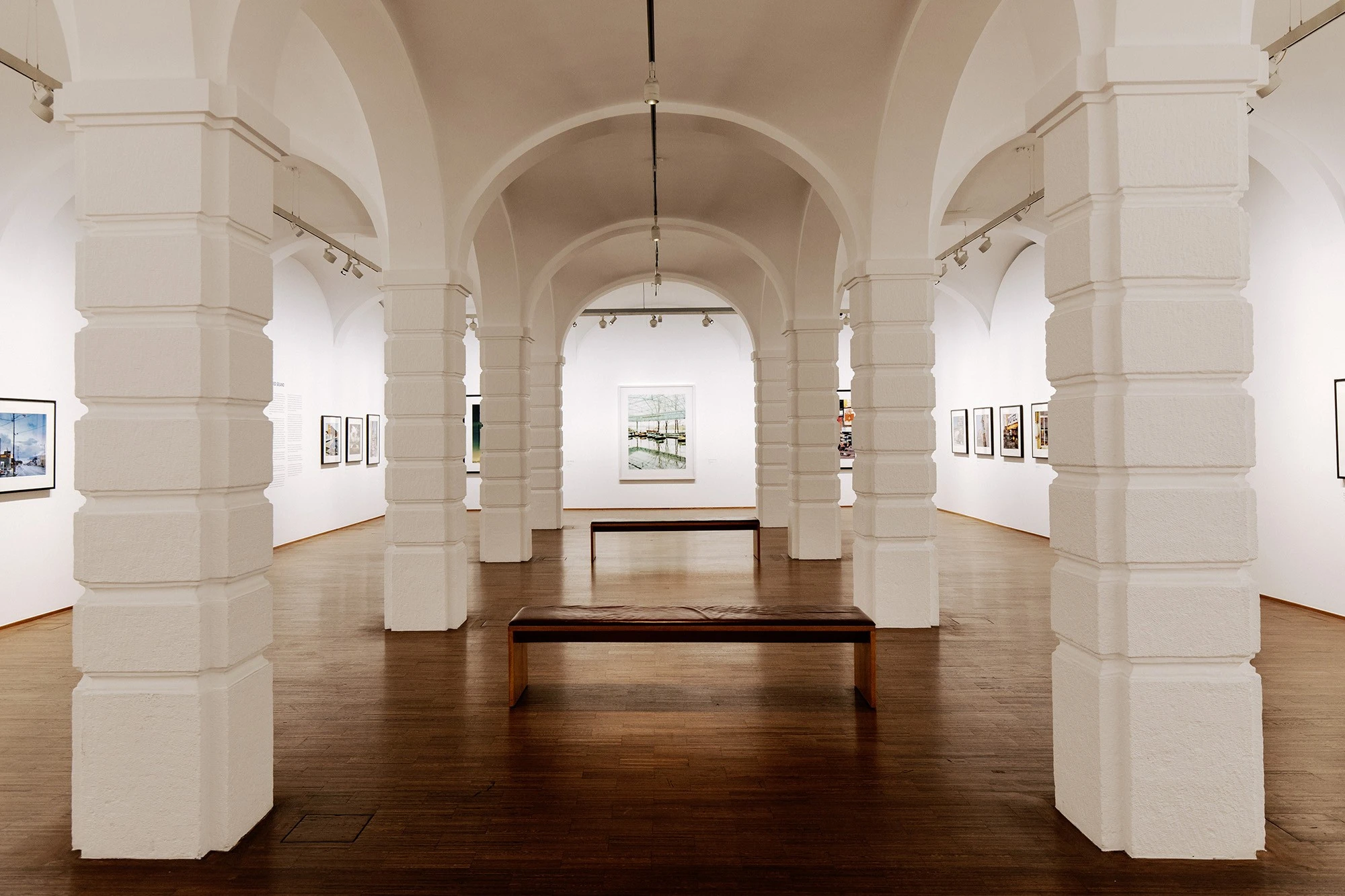 Heller Galerieraum mit weißem Kreuzgewölbe, rhythmisch angeordneten massiven Pfeilern, hölzernem Boden und modernen Fotografien oder Grafiken an den Wänden; in der Mitte zwei schlichte Holzbänke für Besucher.