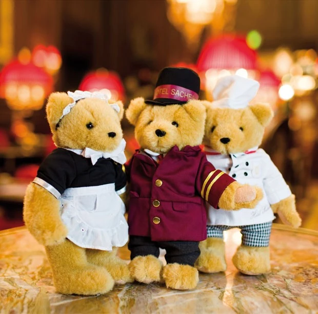 Drei Stoffbären in Hoteluniformen: links als Zimmermädchen mit Schürze und Häubchen, in der Mitte als Page mit rotem „Hotel Sacher“-Outfit und Zylinder, rechts als Koch mit Mütze und gestreifter Hose; posieren gemeinsam auf einem Marmortisch vor warm beleuchtetem Hintergrund.