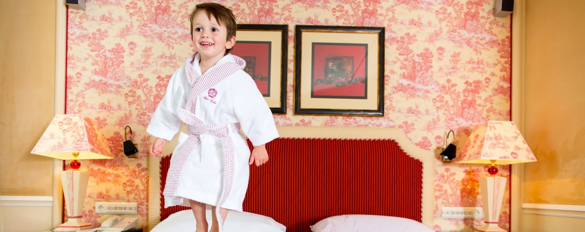 Lächelndes Kind in einem weißen Bademantel mit rot-weiß kariertem Besatz springt auf einem Bett mit rot geripptem Kopfteil, flankiert von zwei Lampen mit passendem Toile-de-Jouy-Muster; im Hintergrund gleichfarbige Tapete und zwei gerahmte Bilder.