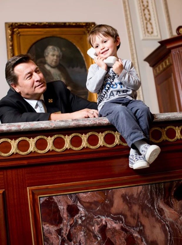 Ein Kind sitzt lachend auf einer marmorverkleideten Hotelrezeption und hält einen Telefonhörer ans Ohr, daneben ein Concierge im dunklen Anzug mit goldenen Schlüsselabzeichen, im Hintergrund ein klassisches Porträtgemälde in prunkvollem Rahmen.