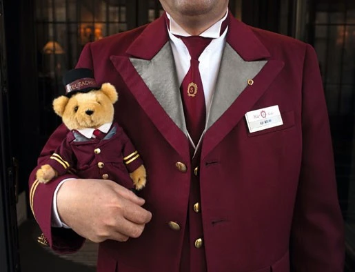 Hotelangestellter in weinroter Uniform mit goldenen Knöpfen und Namensschild, hält einen passend gekleideten Teddybär mit „SACHER“-Mütze im Arm