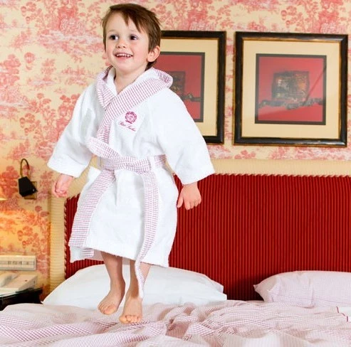 Lächelndes Kind im weißen Bademantel mit rot-weiß karierten Details, springt fröhlich auf einem Bett in einem Zimmer mit rotem Kopfteil, gemusterter Tapete und zwei gerahmten Bildern an der Wand.