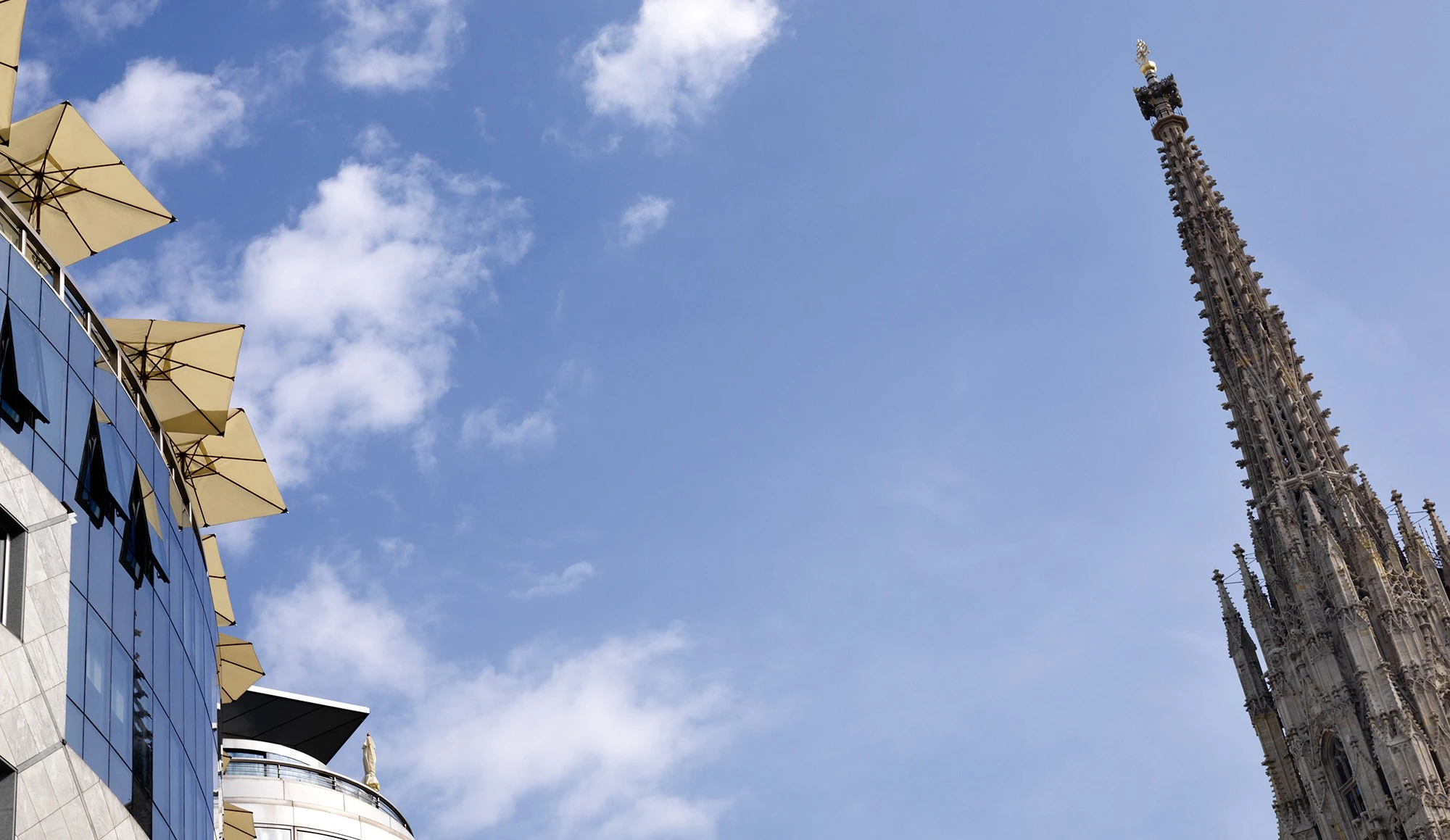 Spitze des Stephansdoms mit filigraner gotischer Architektur ragt rechts ins Bild, links moderne Glasfassade mit gelben Sonnenschirmen auf einer Dachterrasse; blauer Himmel mit vereinzelten Wolken im Hintergrund.