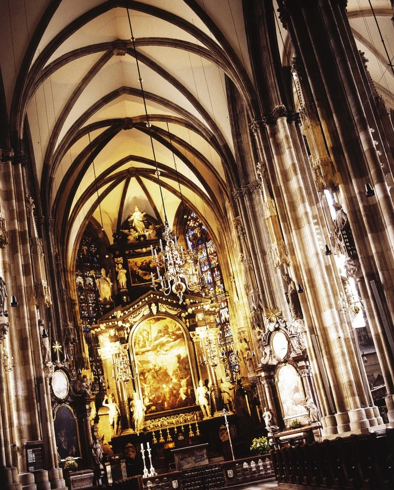 Prunkvoller Innenraum einer gotischen Kathedrale mit hohen Spitzbögen, reich verziertem Altar, vergoldeten Figuren, großen Gemälden und farbigen Glasfenstern; zahlreiche Kronleuchter betonen die vertikale Architektur.